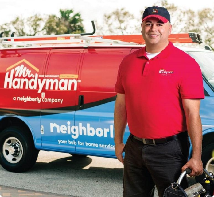 A man in a red shirt and cap stands smiling next to a Mr. Handyman service van, holding tools.