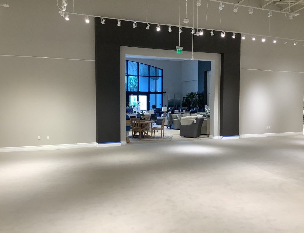 A large, empty showroom with a carpeted floor and overhead lights. In the background, a seating area with tables and chairs is visible through a wide doorway.