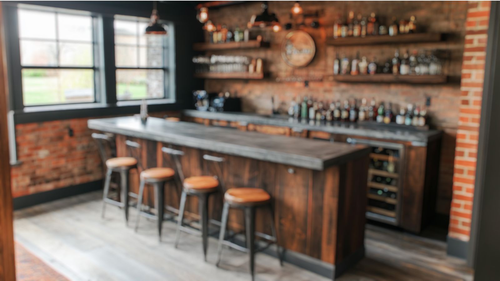 A home bar with a wooden counter, three stools, shelves stocked with bottles, and large windows creates the vibe of a hidden speakeasy in a rustic-style room.